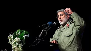 در این تصویر، یک فرد در حال سخنرانی در یک مکان رسمی است. او لباس نظامی به تن دارد و در حال بیان نکاتی با حرکات دست است. در کنار او، میزی با گل‌های تزئینی قرار دارد و میکروفون و بلندگو نیز دیده می‌شود. پس‌زمینه تصویر تاریک است و تمرکز بیشتری بر روی سخنران وجود دارد.