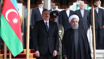 L'image montre deux hommes en costume, probablement des leaders politiques, posant ensemble devant un drapeau azéri. L'un d'eux porte un costume formel, tandis que l'autre est vêtu d'une tenue traditionnelle, avec une robe sombre. À l'arrière-plan, on aperçoit des soldats en uniforme et d'autres personnes, formant une sorte de cérémonie officielle. L'environnement semble être un bâtiment gouvernemental, avec des éléments décoratifs.