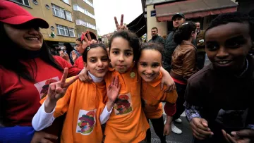 L'image montre un groupe d'enfants souriants, posant ensemble dans une rue animée. Ils portent des t-shirts colorés, principalement orange, avec des motifs ou des logos visibles. Les enfants font des signes de paix avec leurs doigts et affichent des expressions joyeuses. En arrière-plan, on aperçoit des adultes et d'autres participants à un événement festif. L'atmosphère semble conviviale et festive.