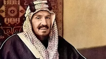 L'image montre un homme assis, portant un habit traditionnel. Il est vêtu d'une thobe sombre et d'un ghutrah, un couvre-chef caractéristique de la culture arabe. Il tient une canne et affiche une expression calme et posée. L'arrière-plan présente un motif décoratif qui renforce le contexte culturel de la scène. Les couleurs de l'image sont enrichies, donnant vie aux vêtements et à l'environnement.