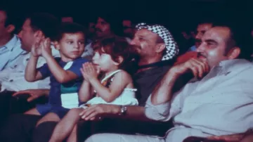 L'image montre un groupe de personnes assises dans une salle, probablement lors d'un événement. Au premier plan, deux enfants, un garçon et une fille, semblent applaudir. Le garçon porte un maillot de bain et la fille est habillée avec une petite robe. Derrière eux, un homme portant un keffieh (le couvre-chef traditionnel) sourit. À sa droite, un autre homme est assis, regardant également vers le devant. L'ambiance paraît festive et conviviale.