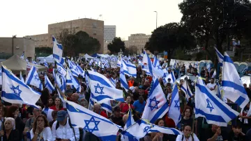 La imagen muestra una multitud de personas en una manifestación, ondeando banderas de Israel. La escena parece vibrante y llena de energía, con una gran cantidad de asistentes que levantan las banderas en un ambiente de protesta o celebración. En el fondo se pueden apreciar árboles y estructuras, lo que sugiere que el evento tiene lugar al aire libre, probablemente en una ciudad. La multitud parece diversa y está unida por una causa común.