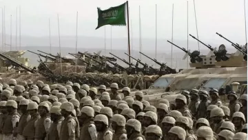 تظهر في الصورة مجموعة من الجنود يرتدون زيًا عسكريًا واقفين في صفوف منتظمة. خلفهم، توجد دبابات ومدرعات عسكرية، حيث تُشاهد المدافع موجهة نحو الأمام. في خلفية الصورة، يوجد علم أخضر يرفرف، مما يوحي بأن هذه الصورة مرتبطة بقوة عسكرية أو استعراض. تعكس الصورة أجواءً عسكرية وهيبة القوات المدافعة.