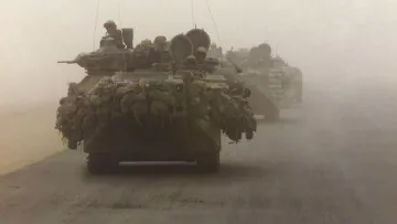 La imagen muestra un convoy militar en un ambiente polvoriento y desértico. Se pueden ver varios vehículos blindados, posiblemente tanques, avanzando en formación. Los vehículos están cargados con equipos y suministros, lo que sugiere que están en una misión de combate o de suministro. La visibilidad es limitada debido a la niebla o polvo, lo que añade una atmósfera tensa a la escena.