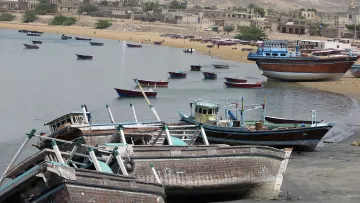 L'image montre une scène côtière avec plusieurs bateaux de pêche échoués sur la plage. On peut voir des petites embarcations peintes en différentes couleurs, certaines abîmées, qui sont disposées le long d'une côte sablonneuse. En arrière-plan, des bâtiments témoignent d'une ville ou d'un village, et le paysage est dominé par des collines ou des falaises. L'eau est calme, et l'atmosphère semble tranquille, avec quelques personnes visibles sur la plage.