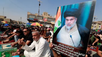 L'image montre une manifestation ou une assemblée publique. De nombreuses personnes sont assises sur des tapis, probablement pour un événement religieux ou politique. Un homme tient une grande affiche avec une photo d'un leader charismatique, visiblement respecté par les participants. Les drapeaux irakiens et d'autres décorations sont présents, ajoutant une ambiance colorée à la scène. Les participants semblent engagés dans une activité collective, reflétant un fort sentiment de communauté et de solidarité.