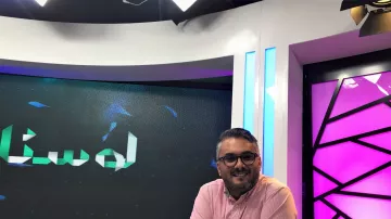 L'immagine mostra un uomo sorridente seduto davanti a una scrivania in uno studio televisivo. Indossa una camicia rosa e presenta un aspetto rilassato e accogliente. Dietro di lui si possono notare schermi che mostrano un logo o un testo in arabo, mentre l'illuminazione dello studio è accentuata da luci colorate, in particolare tonalità di viola e blu. L'ambiente sembra moderno e professionale, tipico di una trasmissione televisiva.