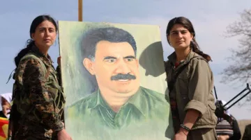 L'image montre deux femmes en tenue militaire tenant un portrait d'un homme avec une moustache. Elles semblent participer à un événement ou une manifestation, entourées d'une atmosphère qui indique une forme de dévotion ou de commémoration. Le fond est naturel, avec un ciel partiellement nuageux. Les expressions des femmes suggèrent détermination et fierté.