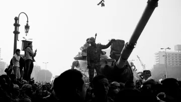 تظهر الصورة مشهداً من مظاهرة أو حدث احتجاجي، حيث يوجد عدد كبير من الناس يتجمعون حول دبابة. أحد الجنود يقف فوق الدبابة ويرفع يدَه كما لو كان يحمل شيئاً أو يجذب الانتباه. الأجواء تشير إلى توتر أو حالة من الحماس بين الحشود. الصورة تُظهر تبايناً كبيراً بين الأضواء والظلال، مما يضفي عليها طابعاً دراماتيكياً.