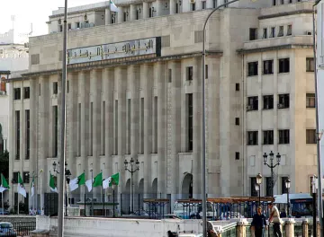 L'image montre un bâtiment majestueux avec une architecture moderne. La façade est claire, avec de grandes fenêtres et des lignes géométriques. On peut également voir des drapeaux algériens flottant devant le bâtiment. Il y a des lampadaires et une ambiance urbaine. Ce contraste entre le style architectural et le décor environnant suggère un lieu important, probablement lié aux institutions gouvernementales.