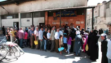 L'image montre une longue file de personnes attendant devant un bâtiment, probablement pour accéder à des services ou des ressources. On peut voir des hommes et des femmes de différentes générations, certains portant des sacs. L'atmosphère semble animée, et le bâtiment est simple, avec une devanture marquée par une enseigne. Des vélos sont stationnés à proximité, et l'environnement urbain est visible en arrière-plan.