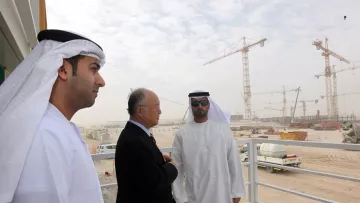 L'image montre trois hommes, apparemment en discussion, sur un balcon ou une plateforme. Ils sont vêtus de vêtements traditionnels des Émirats, comprenant des kandouras et des ghutras. En arrière-plan, on peut voir un site de construction avec des grues et des véhicules, suggérant un projet de développement en cours. L'environnement est poussiéreux et légèrement brumeux, typique des zones de construction dans des climats chauds. Les hommes semblent sérieux et engagés dans leur conversation.
