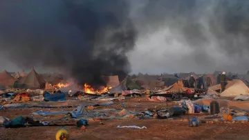 L'image montre un camp de tentes dans un environnement chaotique. On peut voir de la fumée noire s'élever dans le ciel, accompagnée de flammes, indiquant un incendie. Le sol est encombré de débris et de couvertures abandonnées, ce qui suggère une situation difficile. En arrière-plan, des personnes sont visibles, probablement des secouristes ou des autorités, tentant de gérer la situation. L'atmosphère est sombre et perturbée, reflétant une scène de crise ou de catastrophe.