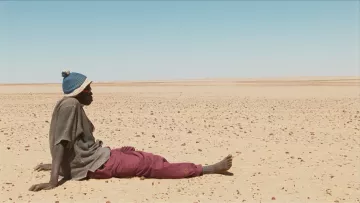L'image montre un homme assis sur le sol désertique. Il porte un pull avec un bonnet et un pantalon de couleur prune. Le paysage autour de lui est vaste et aride, constitué principalement de sable et de petites pierres, s'étendant à perte de vue sous un ciel clair. L'atmosphère semble à la fois isolée et silencieuse, évoquant une sensation de solitude dans cet environnement désertique.
