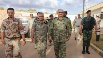 L'image montre un groupe de militaires marchant ensemble dans un environnement urbain. Deux hommes en avant-plan semblent discuter tout en marchant côte à côte. L'un d'eux porte un uniforme camouflage de couleur verte, tandis que l'autre a un uniforme militaire gris avec des distinctions. En arrière-plan, on peut voir d'autres soldats et des véhicules militaires, ce qui indique qu'ils se trouvent dans une zone potentiellement sécurisée. Les soldats semblent concentrés et en mission.