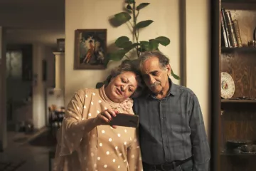 L'image montre un couple âgé, se tenant l'un contre l'autre avec affection. Ils regardent un téléphone ensemble, semblant apprécier ce qu'ils voient. La femme porte une blouse à pois et a un sourire joyeux, tandis que l'homme, en chemise à carreaux, affiche une expression douce. En arrière-plan, on aperçoit un intérieur chaleureux avec des meubles en bois et une plante verte, ce qui crée une ambiance conviviale et intime.