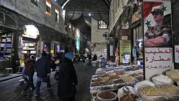 L'image montre une scène d'un souk ou d'un marché typique, probablement en Syrie. On aperçoit des étals avec des épices, des fruits secs, et d'autres produits en vente. Les murs de la rue sont en pierre et semblent anciens, créant une ambiance historique. Des personnes, dont certaines portent des vêtements traditionnels, se déplacent dans la rue, tandis qu'une affiche avec un symbole militaire est accrochée à un mur, ajoutant un élément de contexte culturel et politique à la scène. La lumière faible et les ombres accentuent l'ambiance conviviale et animée du marché.