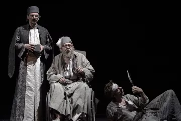 L'image représente une scène de théâtre mettant en avant trois personnages. Deux d'entre eux, habillés de façon traditionnelle et avec des vêtements sombres et ornés, semblent être en position d'autorité ou d'observation. Le troisième personnage est assis dans un fauteuil roulant, avec des vêtements plus clairs et un air pensif ou mélancolique. Il a une longue barbe grise et des lunettes. À côté de lui, un autre personnage, qui paraît être en détresse ou blessé, est au sol, tenant un objet pointu. L'éclairage est sombre, créant une atmosphère dramatique et intense.