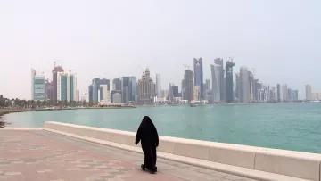 L'image montre une personne vêtue d'une tenue traditionnelle noire, marchant le long d'une promenade au bord de l'eau. En arrière-plan, on aperçoit une ligne d'horizon urbaine avec des gratte-ciel modernes et des bâtiments en construction, représentant une ville côtière. L'ambiance est calme, avec un ciel légèrement brumeux et une mer tranquille. Les pavés de la promenade ajoutent une touche de couleur à la scène.