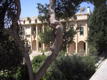 L'image montre un bâtiment en pierre, typique de l'architecture méditerranéenne, avec plusieurs arches et fenêtres. Il est entouré de verdure, avec des arbres et des plantes bien entretenues. Au fond, on remarque un satellite et un ciel bleu clair, ce qui suggère un climat ensoleillé. L'ensemble dégage une atmosphère paisible et accueillante.