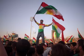 L'image montre une grande foule célébrant, probablement lors d'un événement culturel ou politique. Au premier plan, un homme se tient debout sur une autre personne, tenant des drapeaux kurdes. Il porte un maillot aux couleurs du drapeau kurde, et son expression semble joyeuse et enthousiaste. En arrière-plan, on peut voir de nombreuses autres personnes agitant également des drapeaux, créant une atmosphère festive et énergétique. Le ciel est clair, ce qui ajoute à l'ambiance dégagée de la scène.
