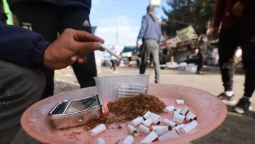 L'immagine mostra una persona che tiene in mano una sigaretta su un piatto di plastica rosa, sul quale sono visibili numerosi mozziconi di sigaretta. Accanto ai mozziconi c'è del tabacco sfuso e un contenitore trasparente. Lo sfondo rivela una strada affollata, con persone che si muovono e diversi cartelloni pubblicitari. L'atmosfera sembra quella di un mercato o di una zona urbana.