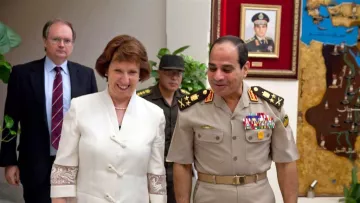 L'image montre une scène de rencontre entre plusieurs personnes dans un environnement officiel. Une femme souriante, vêtue d'une tenue claire, semble discuter avec un homme en uniforme militaire. Ce dernier porte des médailles et des insignias sur son uniforme. En arrière-plan, on peut voir un homme en tenue militaire additionnelle et une carte ou un tableau accroché au mur. L'ambiance semble amicale et détendue, suggérant un dialogue diplomatique ou militaire.