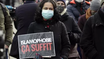 تظهر في الصورة مجموعة من الأشخاص في تجمع عام، حيث تحمل إحدى النساء لافتة مكتوب عليها "ISLAMOPHOBIE ÇA SUFFIT" (الإسلاموفوبيا كفى) باللغة الفرنسية. ترتدي المرأة كمامة وتبدو جادة في التعبير عن قضيتها. الخلفية تحتوي على عدد من الأشخاص الآخرين يرتدون أيضاً الكمامات. تعكس الصورة مشاعر احتجاج ومطالبة بإنهاء التمييز ضد المسلمين.