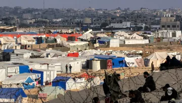L'immagine rappresenta un campo profughi, con numerosi tendoni di vari colori, come blu, bianco e arancione, disposti in un'area sabbiosa. Sullo sfondo si intravedono edifici e una città, mentre una rete di recinzione sembra delimitare il campo. In primo piano, si possono vedere alcune persone sedute, creando un senso di comunità nonostante le difficili condizioni di vita. La scena evoca una forte emozione, evidenziando la realtà delle persone costrette a vivere in tale situazione.