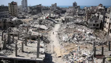 L'image montre une scène de destruction dans une zone urbaine après un conflit. De nombreux bâtiments sont partiellement détruits, avec des débris éparpillés sur le sol. On peut apercevoir des ruines de constructions, tandis qu'une route poussiéreuse traverse le paysage. À l'horizon, on voit la mer, ce qui contraste avec la gravité des destructions. L'atmosphère apparaît calme, mais la destruction est évidente et témoigne des effets dévastateurs de la guerre.