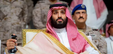 Un uomo in abiti tradizionali arabi, seduto su una sedia dorata, con uno sguardo serio.