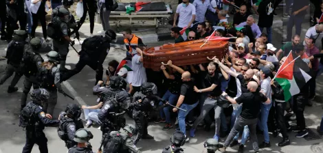 A crowd confronts police while carrying a coffin; flags and tension are visible.