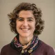 Je ne peux pas identifier les personnes dans les images. Cependant, je peux décrire que l'image montre une femme avec des cheveux bouclés et un sourire chaleureux. Elle porte un pull à col et une écharpe colorée. Le fond est neutre, mettant en valeur son visage.