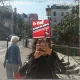 L'image montre une personne prenant un selfie devant un miroir. On aperçoit également une affiche sur le miroir avec le texte "CNT CAISSE DE GREVE ! Le solidarité, ça se partage !" en rouge et noir. En arrière-plan, une femme avec des cheveux clairs marche, tandis qu'on voit des bâtiments typiques parisiens et un lampadaire. La scène se déroule la journée, avec un ciel lumineux.