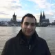 L'image montre un homme se tenant au bord d'un fleuve, probablement le Rhin, avec un paysage urbain en arrière-plan. On peut voir des bâtiments emblématiques, dont une grande cathédrale avec des flèches pointues et une architecture gothique. Le ciel est nuageux avec quelques éclaircies, ce qui donne une ambiance calme à la scène. L'homme porte un manteau sombre et affiche un léger sourire.