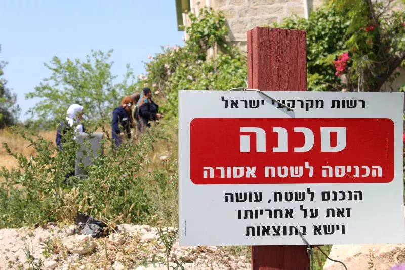 L'image montre un panneau en hébreu, avec le mot "סכנה" (danger) mis en évidence. Le panneau indique des instructions ou des avertissements concernant l'entrée dans une zone spécifique. En arrière-plan, on peut apercevoir des personnes qui semblent être occupées à une activité dans la nature, entourées de végétation. L'environnement semble naturel, avec des arbres et des buissons.