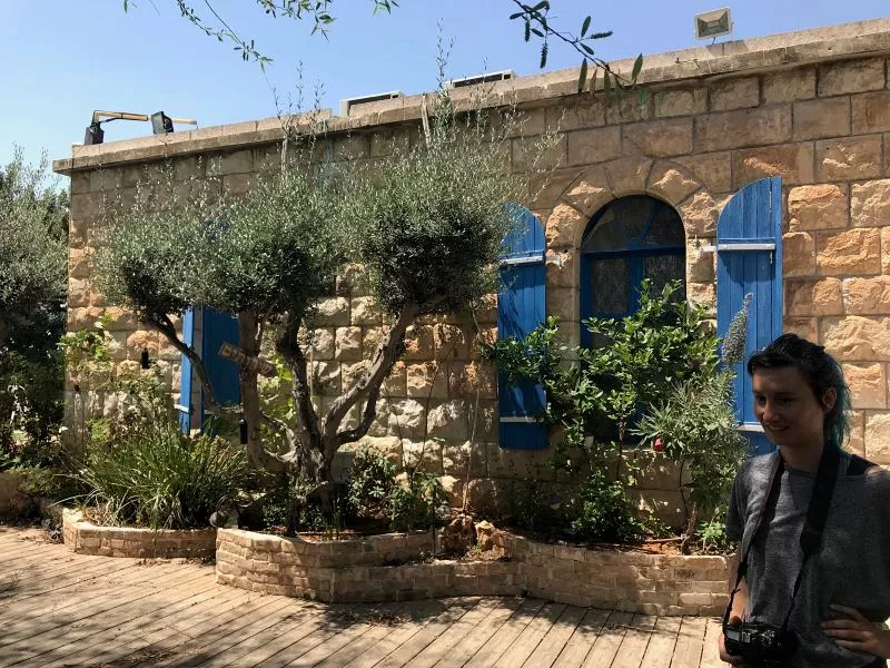 L'image montre un mur en pierre d'une maison avec des volets bleus. Il y a de petites plantes et des arbres autour de la maison, ajoutant une touche de verdure au paysage. Sur le côté droit, une personne se tient près de la maison, tenant un appareil photo. Le ciel est clair et ensoleillé, créant une atmosphère chaleureuse et accueillante.