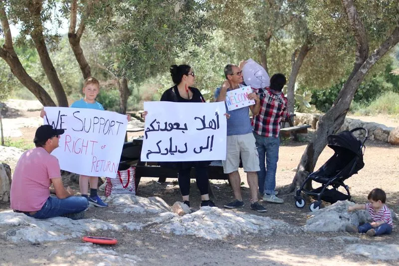 L'image montre un groupe de personnes en train de manifester dans un espace extérieur, probablement un parc. Certains participants tiennent des pancartes avec des messages en anglais et en arabe. Un enfant blond se trouve parmi eux, tandis qu'une femme tient une grande pancarte. Au fond, on aperçoit d'autres personnes, dont un enfant assis près d'une poussette. L'ambiance semble être celle d'une réunion pacifique pour exprimer un soutien ou des revendications.