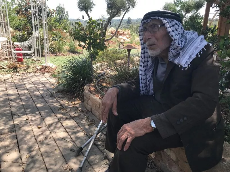 L'image montre un homme âgé assis sur un chemin en bois dans un jardin ou un espace extérieur. Il porte un habit traditionnel avec un keffieh sur la tête, et il utilise une canne pour se soutenir. L'environnement est verdoyant, avec des plantes et des arbres en arrière-plan. On peut sentir une atmosphère paisible et contemplative.
