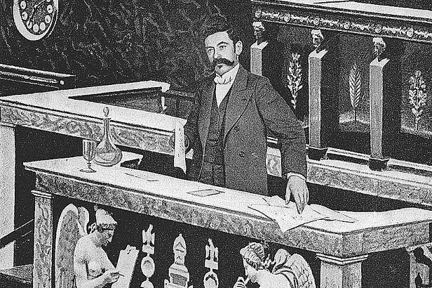 L'image montre un homme bien habillé, debout derrière un bureau ou une table qui semble faire partie d'une institution, probablement un parlement ou une assemblée. Le bureau est décoré avec des motifs ornementaux et des sculptures d'anges de chaque côté. L'homme semble concentré, tenant des documents, et l'ambiance générale est formelle et historique.
