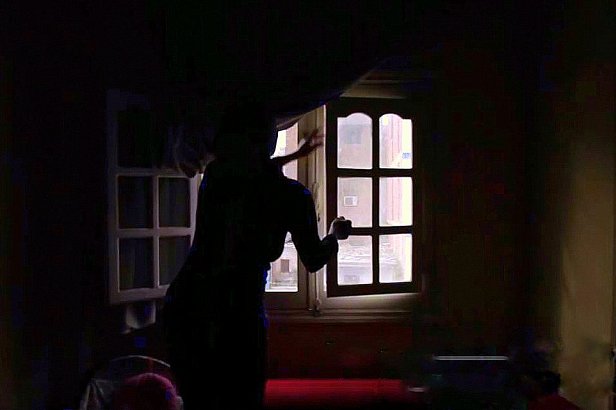 L'image montre une silhouette se tenant près d'une fenêtre dans une pièce sombre. La lumière entre par la fenêtre, illuminant partiellement l'espace environnant, tandis que des détails tels que les murs et les meubles sont à peine visibles dans l'ombre. La silhouette semble en mouvement, ajoutant une ambiance dynamique et mystérieuse à la scène. Les rideaux sont légèrement ouverts, laissant entrevoir l'extérieur.
