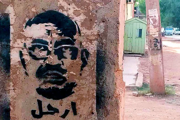 L'image montre un graffiti sur un mur, représentant un visage stylisé d'un homme avec des lunettes. Le dessin est en noir, sur un fond de béton clair. En arabe, le mot "ارحل" est inscrit, ce qui signifie "pars" ou "sors". L'environnement semble urbain, avec des bâtiments et une route visible en arrière-plan.