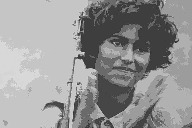 L'image montre une femme avec des cheveux bouclés, tenant un fusil. Elle sourit légèrement tout en applaudissant, ce qui semble indiquer un moment de célébration ou de camaraderie. Son uniforme militaire est visible, ce qui suggère qu'elle est impliquée dans un contexte de lutte ou de conflit. L'arrière-plan est flou, mettant l'accent sur sa figure.