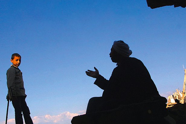 L'image montre deux silhouettes. À gauche, un garçon se tient debout, regardant vers l'autre personne. À droite, un homme est assis, en train de parler, ses bras gesticulant. Le fond est un ciel bleu, ce qui crée un fort contraste avec les silhouettes noires. L'environnement semble brut, avec des débris visibles, indiquant un cadre peut-être urbain ou une zone touchée par des événements difficiles.