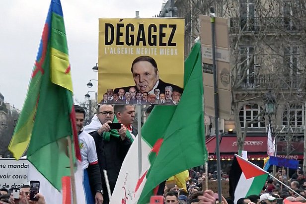 L'image montre une manifestation avec des participants portant des drapeaux. Au centre, un homme tient une grande pancarte sur laquelle est écrit "Dégagez l'Algérie mérite mieux", accompagnée d'une photo d'une personne. Les manifestants semblent exprimer des opinions politiques, et l'atmosphère semble être celle d'une contestation pacifique. Des slogans sur les pancartes reflètent des revendications pour des changements en Algérie.