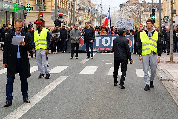 L'image montre une manifestation dans une rue où un groupe de personnes tient une grande banderole avec des slogans. Au premier plan, deux agents de sécurité ou policiers portent des gilets réfléchissants et semblent surveiller la situation. Des manifestants sont visibles à l'arrière, et il y a une ambiance de mobilisation avec des drapeaux. L'environnement urbain se présente avec des bâtiments voisins et une circulation routière réduite à ce moment.