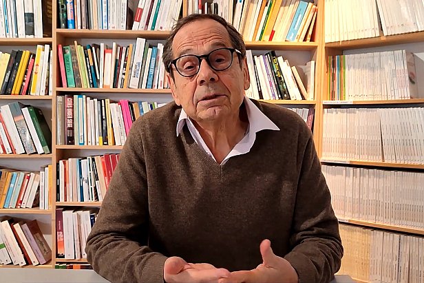 L'image montre un homme assis à une table, s'exprimant avec des mains gesticulantes. Il porte un pull brun et une chemise blanche. Derrière lui, une bibliothèque bien fournie avec des livres de différentes tailles et couleurs crée un environnement studieux et accueillant. L'homme semble être en train de partager des idées ou des réflexions.