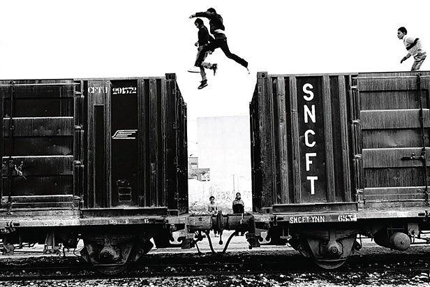L'image montre un groupe de jeunes qui sautent d'un wagon de train à un autre. La scène est en noir et blanc, ce qui accentue l'action dynamique et l'intensité du moment. On peut observer deux jeunes sautant dans les airs, tandis que d'autres se trouvent sur les wagons ou à proximité. L'environnement semble urbaine, avec une atmosphère à la fois aventureuse et un peu risquée. Les détails des wagons et l'arrière-plan renforcent l'impression d'un cadre industriel.